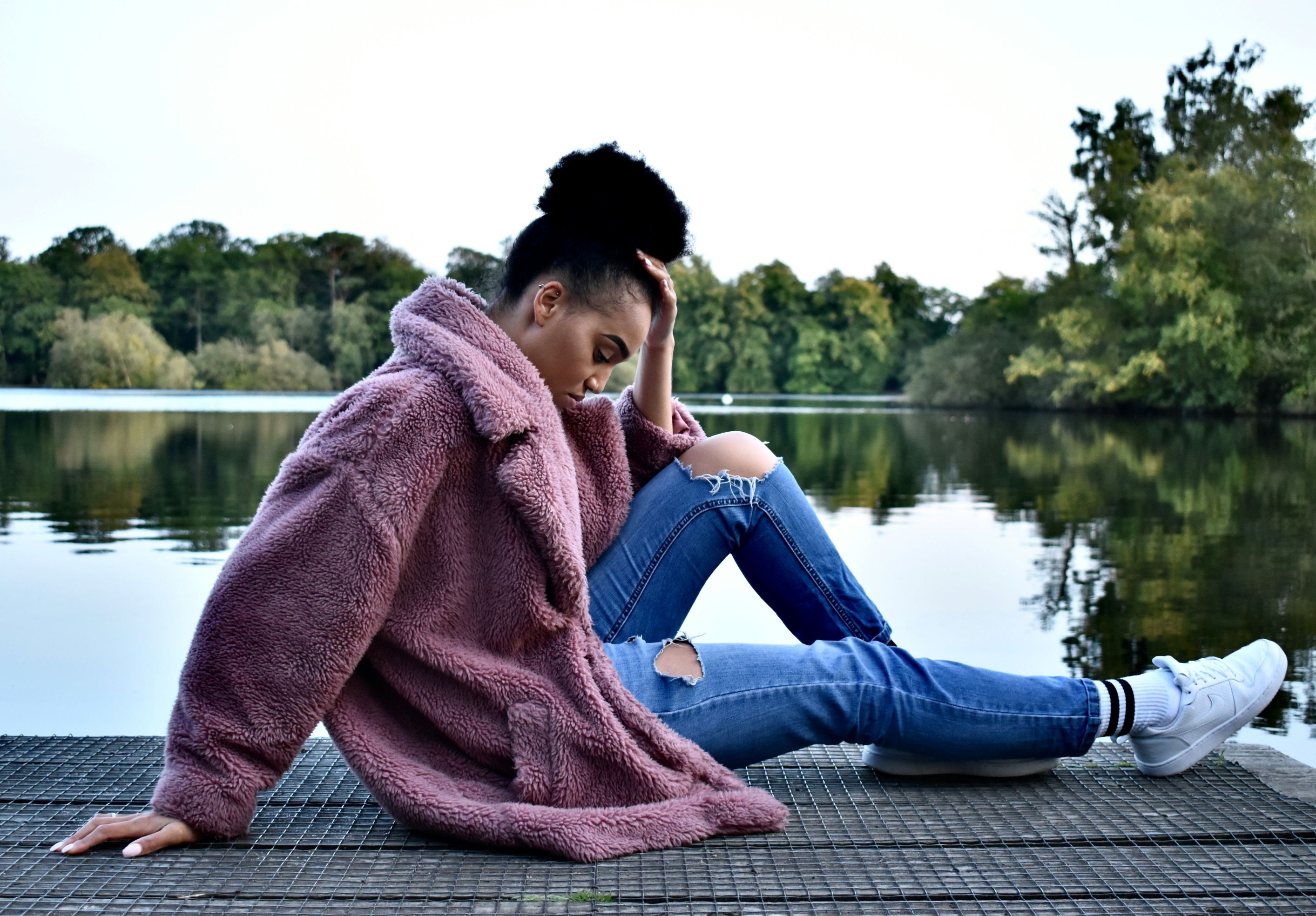 A photograph of a woman wearing a pink jacket and distressed blue jeans sitting on wooden dock during daytime