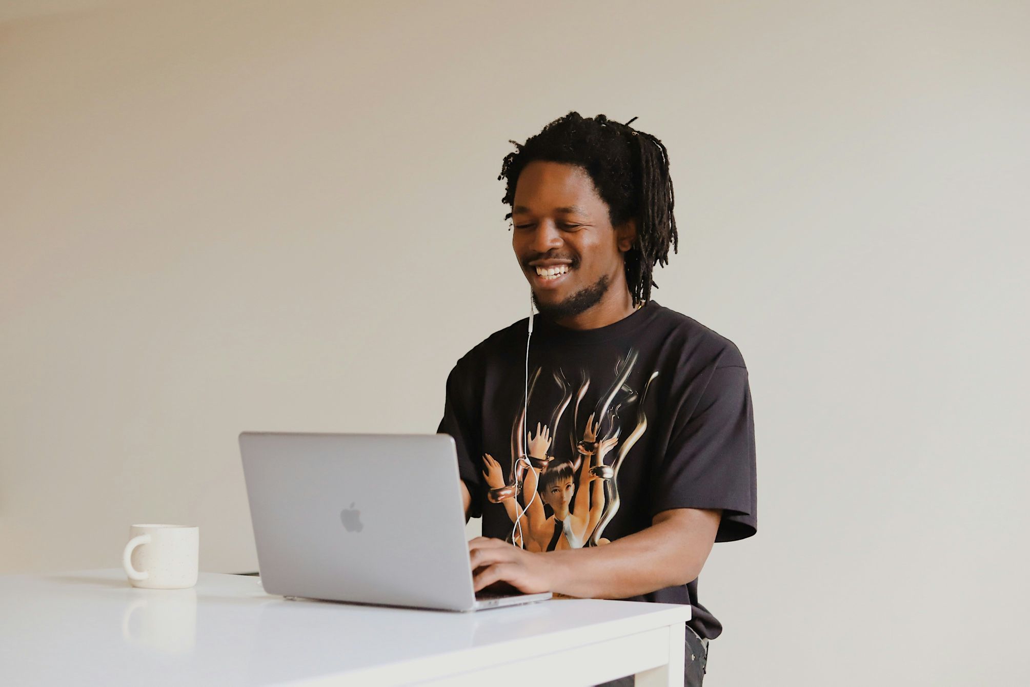 A photograph of a man wearing a black t-shirt sitting at a table smiling at laptop