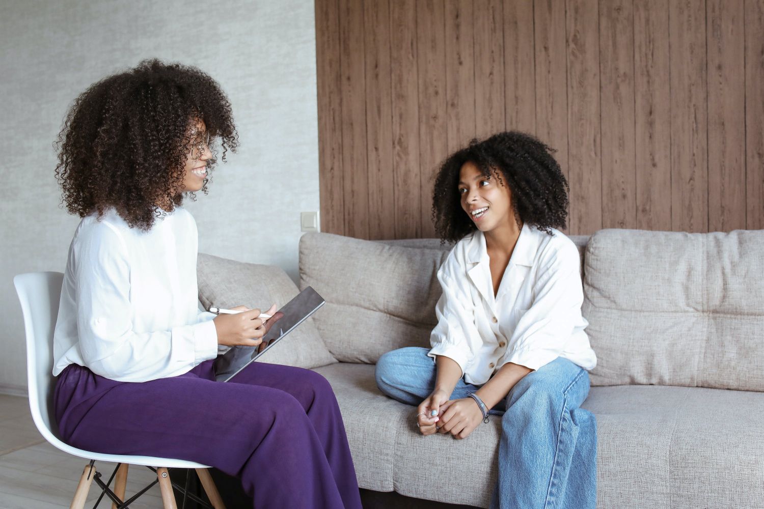 A photograph of a woman sitting on a couch talking to another woman sitting in a chair.