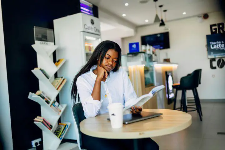 Woman wearing a white button up shirt resting her elbow on a small round table and her chin on her knuckle. THere is a laptop and coffee cup on the table. Woman is reading a booking in a cafe.