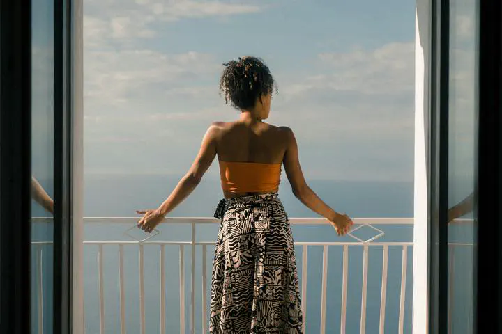 A woman wearing an orange top and multicolored skirt standing on a balcony with her back turned facing the water.