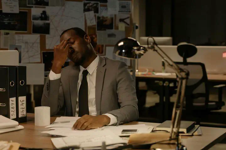 a Black man wearing a suit sitting at a desk with his hand to his face and his eyes closed.